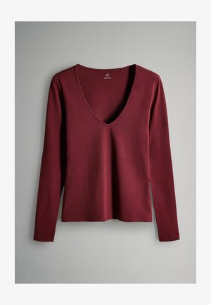 Dlouhý rukáv hluboce červené tričko s V výstřihem leží na šedém povrchu, vyrobené z hladké bavlněné tkaniny.