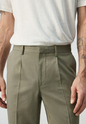 Uomo che indossa pantaloni sartoriali verde oliva e una camicia beige chiaro a maniche corte infilata, con tatuaggi visibili sugli avambracci.