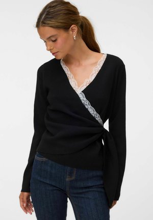 Frau mit braunen Haaren im Pferdeschwanz trägt einen schwarzen Wickelpullover mit weißer Spitzenborte und dunkelblaue Jeans, schaut nach unten zu ihrer linken Seite.