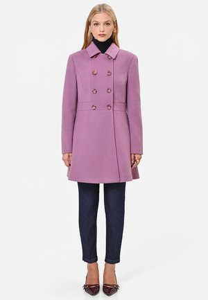 Donna che indossa un cappotto viola doppiopetto, dolcevita nero, jeans blu scuro e décolleté bordeaux con punta, in piedi su sfondo bianco.