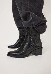 Botas de tobillo de cuero negro con relieve de cocodrilo, con punta afilada y tacón cuadrado, que presentan una banda elástica lateral y una superficie lisa y texturizada.