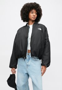 Czarna krótka kurtka bomberka z zamkiem z przodu, ściągaczami na rękawach, delikatnym połyskiem oraz logo The North Face na lewej piersi; zestawiona z jasnoniebieskimi jeansami.