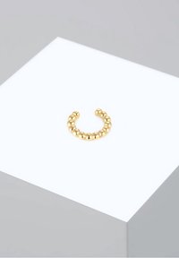 Bague ouverte en perles dorées présentée sur un piédestal géométrique blanc sur fond gris clair.