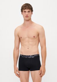 Schwarze Boxer-Shorts mit einem grauen elastischen Bund, der das "HUGO"-Logo zeigt. Das Material wirkt glatt, mit einem engen Schnitt und minimaler Naht.