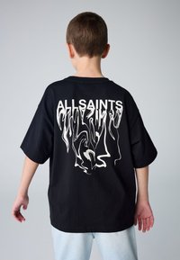Laps vaadatuna tagantpoolt, seljas must, liiga suur T-särk moonutatud valge "ALLSAINTS" kirjaga ning helesinised teksad, taustaks hall sein.