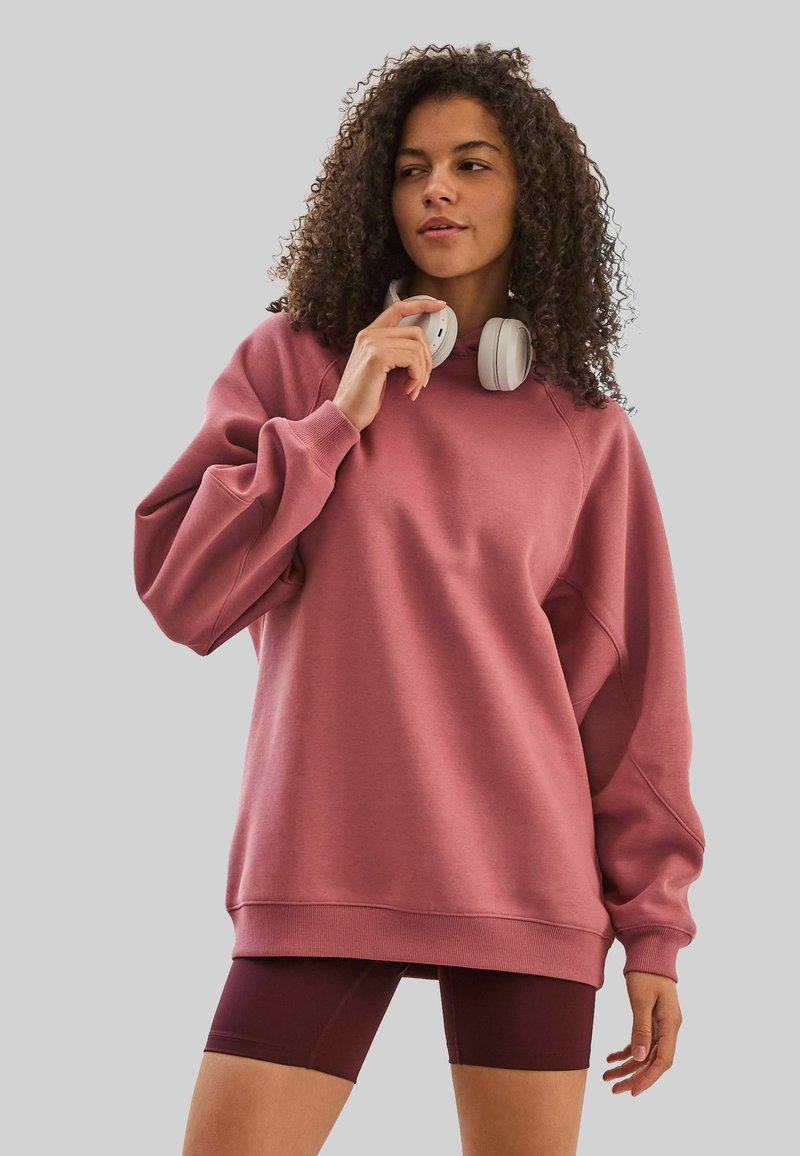 Sweatshirt oversize rose clair avec un col rond, des poignets et un ourlet côtelés. Associé à un short bordeaux ajusté. Casque audio blanc porté autour du cou.