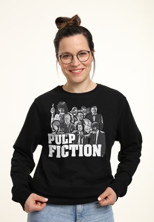 Kvinde med briller og hårknytte bærer sort sweatshirt med Pulp Fiction-figurer og titel i hvid tekst.