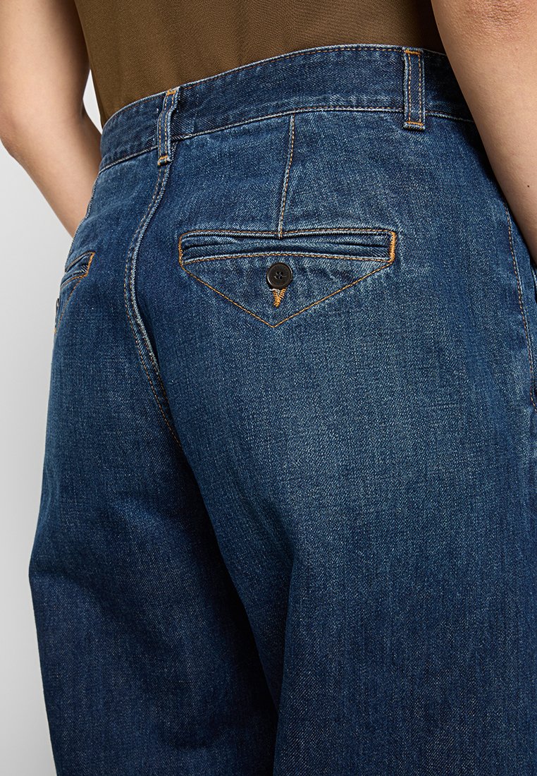 Ciemnoniebieskie dżinsy z wysokim stanem, z tylną kieszenią o trójkątnym wzorze, zapinaną na jeden czarny guzik i pomarańczowe szwy.
