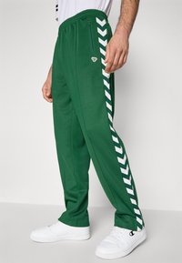 Zöld futónadrágot viselő férfi fehér sorakozó csíkokkal az oldalán, cipzáros zsebbel és fehér tornacipővel, egyszínű háttér előtt.