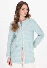Giacca in tweed azzurro chiaro con texture, colletto rotondo, tasche con bottone, bordi frangiati e taglio dritto. Dettagli sottili lungo le cuciture.