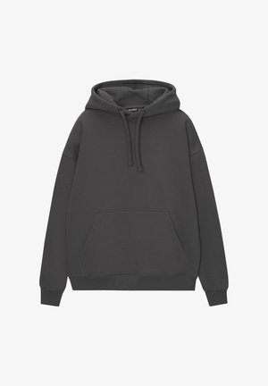 Persona con vaqueros azules descoloridos de gran tamaño, sudadera con capucha gris oscuro con las manos en el bolsillo delantero y zapatillas marrón oscuro sobre fondo blanco.