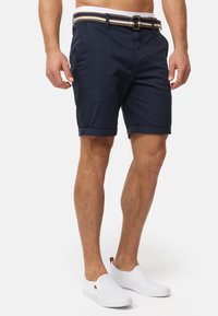 Navy Shorts aus Baumwollstoff mit umgeschlagenem Saum, ausgestattet mit einem kontrastierenden gestreiften Gürtel und Seitentaschen. Tragen mit weißen Slippern.