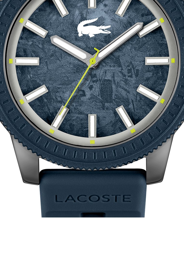 Montre Lacoste bleue avec un cadran texturé, des index horaires blancs, et une aiguille des secondes verte vif, équipée d'un bracelet en silicone et d'une lunette nervurée.