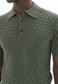 Olíva zöld kötött pólóing texturált mintával. Klasszikus gallérral, három barna gombbal és rövid ujjakkal rendelkezik.