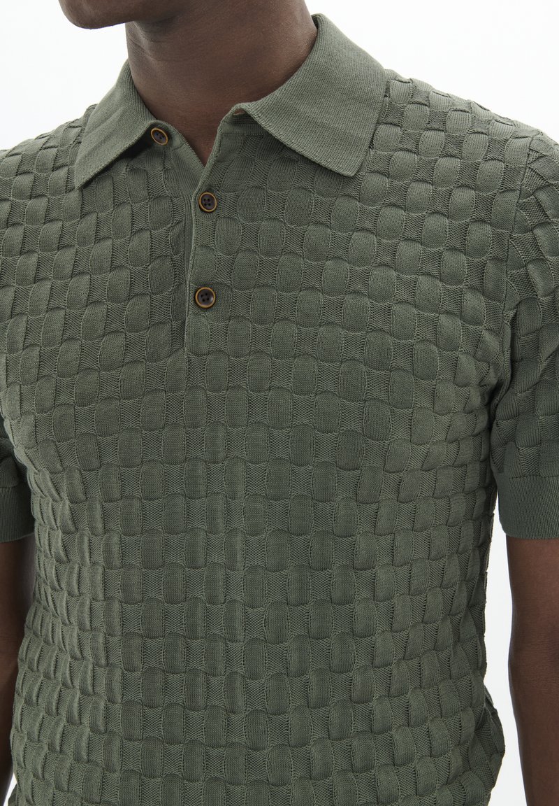 Strick-Poloshirt in Olivgrün mit einem strukturierten Muster. Verfügt über einen klassischen Kragen, drei braune Knöpfe und kurze Ärmel.