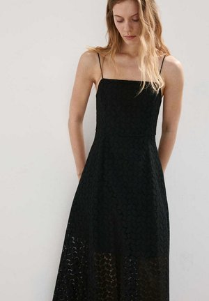 Jeune femme aux longs cheveux blonds portant une robe noire en dentelle sans manches avec de fines bretelles, regardant vers le bas sur un fond uni.