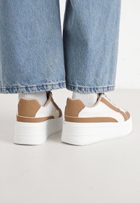 Baskets blanches et marron avec un dessus texturé, semelle plateforme et accents contrastants. Portées avec un pantalon en denim large de couleur bleu clair.