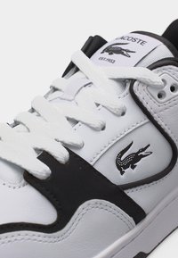 Λευκό και μαύρο αθλητικό παπούτσι Lacoste με λευκά κορδόνια και ορατό λογότυπο κροκόδειλου στη πλευρά και στη γλώσσα, κοντινή λήψη.