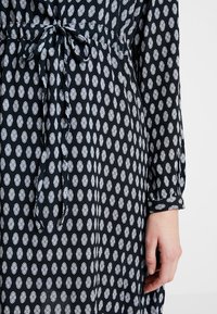 Robe noire avec des motifs ovales blancs, cintrée à la taille avec un lien. Dispose de manches longues avec des poignets retroussés. Texture de tissu douce et fluide.