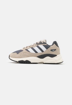 Scarpe da ginnastica beige e blu navy con materiali in suede e mesh, dotate di tre strisce bianche, punta rotonda, suola ammortizzata e dettagli neri.