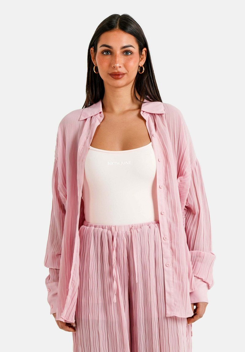 Camisa plisada de color rosa pálido con cuello y botones, superpuesta a una blusa ajustada blanca y pantalones plisados a juego, mostrando un aspecto texturizado.