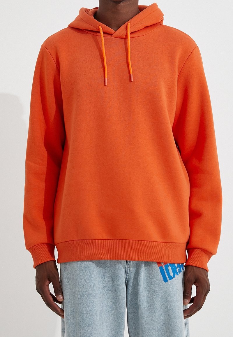 Personne portant un sweat à capuche orange vif avec des cordons assortis et un jean bleu clair avec un graphisme bleu sur la partie supérieure de la cuisse.