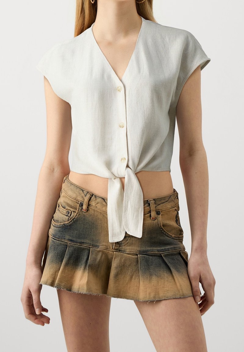 Femme portant un crop top blanc à manches courtes boutonné et noué à la taille, associé à une mini jupe en denim plissée, décolorée brune et noire.
