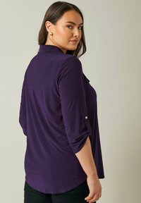 Dark purple, long-sleeve blouse with a collar, rolled sleeves secured by buttons, and a rounded hemline. Smooth, lightweight fabric.