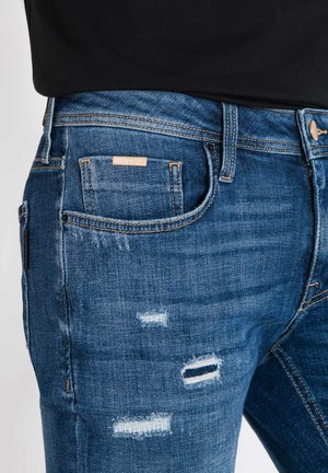 Jeans de mezclilla azul descolorido con detalles rasgados, cinco bolsillos, costuras en contraste y una etiqueta de parche en la cintura trasera.