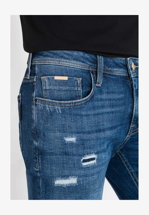 Jean en denim bleu délavé avec détails déchirés, cinq poches, coutures contrastées et une étiquette en patch au dos de la taille.