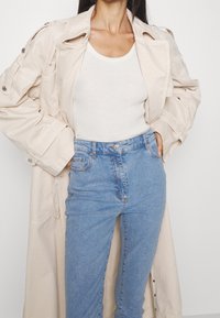 Trench coat beige in cotone sopra un top bianco a costine, abbinato a jeans a vita alta di colore blu chiaro, con il classico design a cinque tasche.