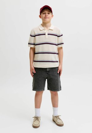 Poiss punase nokamütsiga, kes kannab kreemikat triiksärki lühikeste varrukatega, tumesiniseid lühikesi pükse, valgeid sokke ja beežide jalanõudega, seisab valge tausta ees.