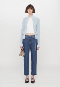 Ανοιχτό μπλε ζακέτα με φερμουάρ και ριγωτή υφή, λευκό cropped top, κατευθείαν μπλε τζιν, και τσάντα με σχέδια. Σκοτεινά, επίπεδα παπούτσια.