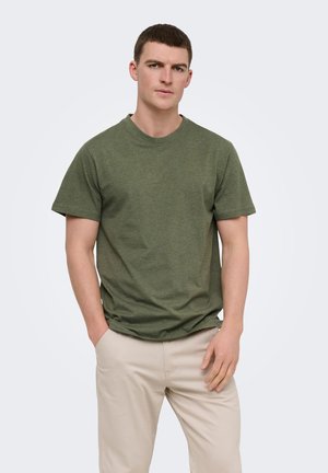 Young man with short brown hair wearing an olive green short-sleeve t-shirt and beige pants, standing against a plain light background.
