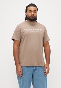 Ruskea puuvillainen t-paita, jossa on lyhyet hihat ja pyöreä kaula-aukko. Edessä valkoinen logopainatus ja rento istuvuus.