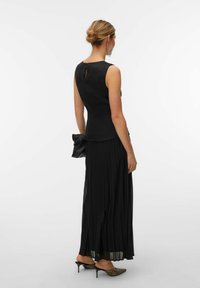 Femme avec un chignon blond portant une robe noire sans manches, tenant une pochette noire et portant des talons aiguilles noirs pointus à talon bas.