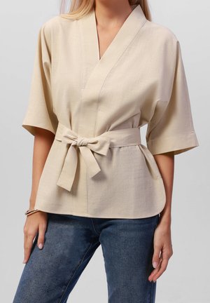 Beige wikkelblouse met korte mouwen, een gestrikte taille, een gladde textuur en een subtiel geweven patroon. Gecombineerd met een blauwe spijkerbroek voor contrast.