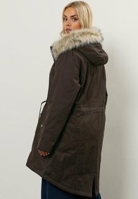 Kvinna med blont hår som bär en mörkbrun parkas med en foder av päls, vänd i sidled mot en enkel ljus bakgrund.