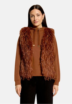 Vrouw met steil donker haar draagt een bruine faux fur-vest over een bruine hoodie en zwarte broek tegen een effen lichte achtergrond.