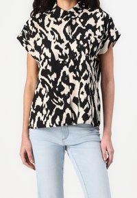 Chemise noire et blanche à manches courtes, présentant un motif abstrait audacieux, une fermeture à boutons sur le devant et un col pointu. Le tissu a une texture lisse.