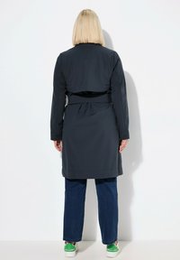 Lungo trench grigio scuro con cintura, pieghe posteriori e maniche lunghe. Abbinato a jeans blu e sneakers bianche con dettagli verdi.