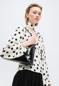 Γυναίκα με λευκό πουκάμισο με μαύρες πουά και αντίστοιχο μαντήλι στο λαιμό, κρατώντας μια μαύρη τσάντα ώμου, ποζάροντας μπροστά σε απλό φόντο.