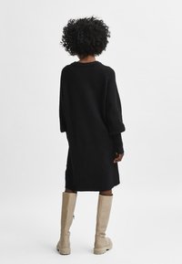 Juodas megztas suknelė laisvo silueto, ilgomis rankovėmis ir vidutinio ilgio apačia, derinama su smėlio spalvos aukštakulniais batais iki kelių. Minkšta faktūra ir paprastas dizainas.