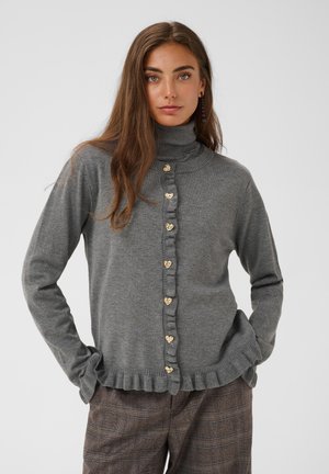 Femme aux longs cheveux bruns portant un pull gris à col roulé avec des boutons en forme de cœur et un bord volanté, associée à un pantalon marron à carreaux.