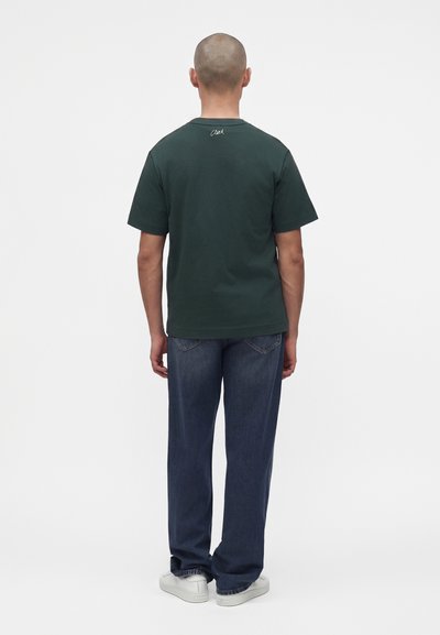 Homme debout, dos tourné vers l'avant, portant un t-shirt vert foncé, un jean bleu et des baskets blanches sur un fond blanc uni.