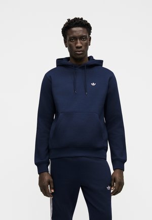 Sort mand iført en marineblå hættetrøje og matchende bukser med hvide Adidas trefoil-logoer, stående mod en ensfarvet lys baggrund.