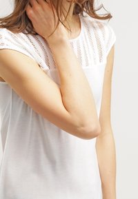 Vit topp med en meshpanel över axlarna och övre bröstet. Mjukt tyg, korta ärmar och en avslappnad passform.