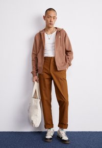 Sudadera marrón con cremallera, camiseta sin mangas blanca, pantalones marrones y zapatillas beige con acentos en azul. Sosteniendo un bolso de crema con diseño azul.