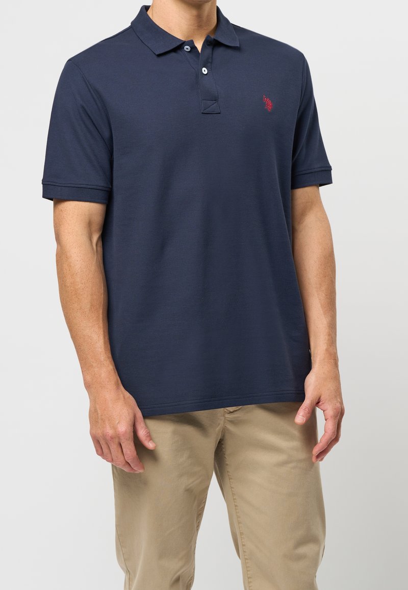 Homme portant un polo bleu marine à manches courtes avec un logo rouge sur la poitrine et un pantalon beige, debout devant un fond clair uni.