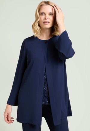 Mujer rubia con cabello ondulado que lleva una chaqueta azul marino de manga larga y abierta sobre un top de lentejuelas, sobre un fondo claro y liso.